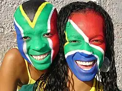 Dos personas con caras pintadas de la bandera de Sudáfrica. Llevan los colores de la bandera: verde, azul, rojo y negro, con líneas blancas y amarillas.
