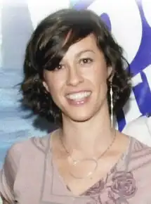 A Caucasian female with light colored hair leaning her head back while singing into a microphone. She is wearing a sleeveless green shirt with dark grey pants and has a guitar strapped around her.