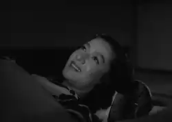 Noriko, vestida con el traje típico, recostada en un futón y cubierta con una manta, mira fijamente al techo y sonríe.