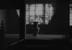 En una habitación semioscurecida, se ve un jarrón ordinario en el suelo al fondo, detrás del cual hay una pantalla sobre la que aparecen las sombras de las ramas.